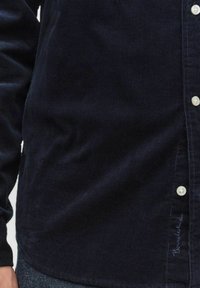 Marinblå kordskjorta med texturerad yta, med knäppning framtill och vita knappar. Diskret broderad logotyp vid fållen.