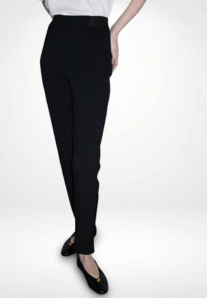 Frau trägt eng anliegende schwarze Hose und schwarze Ballerinas mit goldenen Akzenten, steht mit einem Bein über das andere geschlagen vor einem schlichten Hintergrund.