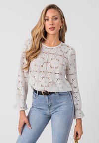 White lace blouse with floral cutouts, round neckline, and long puff sleeves. Paired with blue high-waisted denim jeans and a black belt.