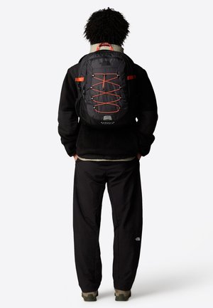 Black backpack with orange accents and bungee cord design, featuring a zipper front, padded straps, and mesh side pockets.