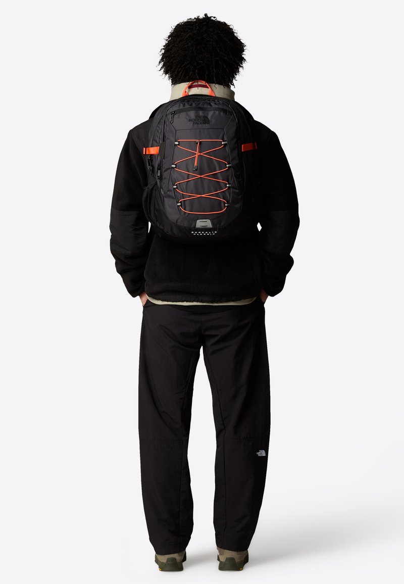 Mochila preta com detalhes laranja e design de corda elástica, apresentando frente com fecho de correr, alças almofadadas e bolsos laterais em malha.