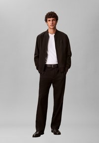 Blouson aviateur noir porté sur une chemise blanche, associé à un pantalon noir et des chaussures noires. Coupe droite et tissu lisse. Détails simples.