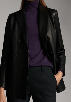 Femme portant un blazer en cuir noir à double boutonnage, un pull à col roulé violet foncé et un pantalon sombre, une main dans la poche.