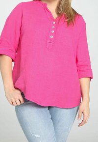 Chemisier en lin rose sans col, avec une patte de boutonnage à cinq boutons et des manches retroussées, associé à un jean bleu clair déchiré.