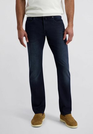 Mann trägt dunkelblaue Jeans, beige Wildleder-Slipper und ein weißes Hemd, steht vor einem einfarbigen grauen Hintergrund.