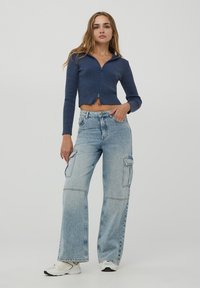 Granatowy sweter crop z prążkowanego materiału, z zapięciem na zamek z przodu i kołnierzem, w połączeniu z szerokimi, jasnoniebieskimi dżinsami cargo z bocznymi kieszeniami i subtelnymi przeszyciami.