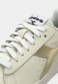 Close-up of beige leather sneaker with white laces and gold diagonal stripe, showing part of the branded tongue label.