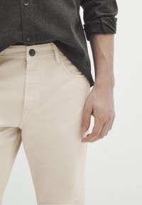 Beige bomullschinos med slim fit. Framknappstängning och sidofickor. Bärs med en mörk strukturerad skjorta, visad vid midjan.