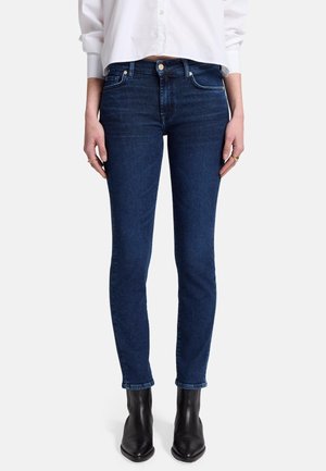 Persoon draagt donkerblauwe slim-fit jeans, wit overhemd en zwarte enkellaarsjes, staat rechtop met de handen ontspannen langs het lichaam.
