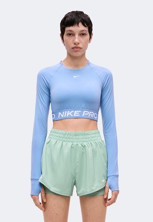 Žena oblečená v svetlomodrý crop top s dlhými rukávmi a mätovo zelené športové šortky stojaca na jednofarebnom pozadí.