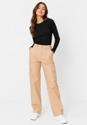Woman standing with hands in pockets wearing black long-sleeve top, beige wide-leg cargo pants, and white sneakers against a plain background.