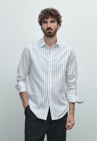 Mann mit lockigem Haar und Bart, der ein weißes, knopfbares Hemd mit grauen vertikalen Streifen und schwarze Hosen trägt, und mit einer Hand in der Tasche steht.