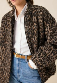 Veste imprimée léopard en brun et noir, avec un zip à l'avant, un col côtelé et une coupe oversized, superposée sur une chemise blanche.