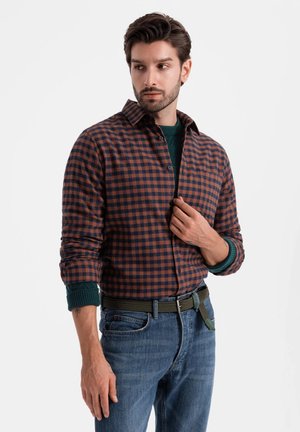 Heren langslovige, geruite shirt in bruin en marineblauw, met een knoopsluiting, kraag en opgerolde mouwen. Draag het over een groene trui en denim jeans.