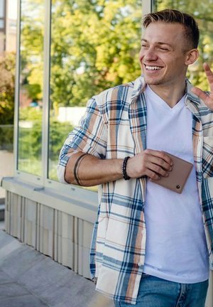 Kostkovaná košile s límečkem v béžové, modré a oranžové barvě přes bílém tričku. Drží světle hnědou peněženku, s viditelným tetováním na zápěstí.