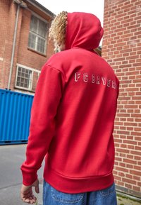 Felpa rossa con cappuccio, con la scritta "FC BAYERN" in lettere bianche sulla schiena. Il tessuto è liscio con polsini e orlo a costine.