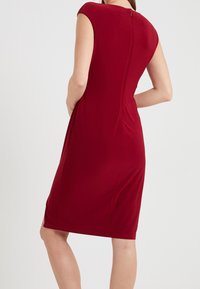 Nainen, jolla on päällä tiukka hihaton polvipituinen punainen mekko, jossa on takavetoketju, seisoo kädet koukistettuina yksinkertaista vaaleaa taustaa vasten.