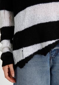 Svetr s černo-bílými pruhy, pletená textura, volný střih, s nezačištěnými okraji a širokými rukávy, nošený přes modré džíny.