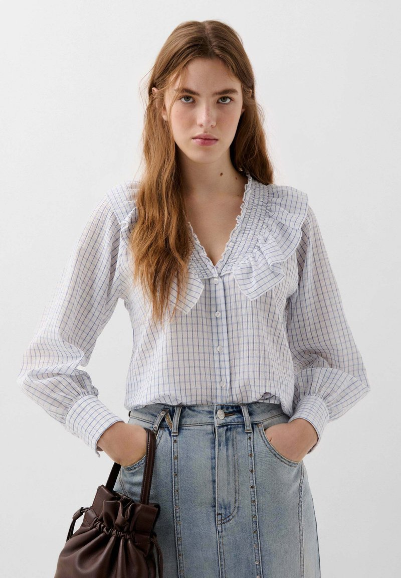 Camicia a quadri blu e bianchi con colletto arricciato e maniche lunghe, abbinata a una gonna di jeans e una borsa a tracolla marrone con cordoncino.