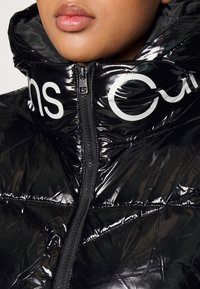 Schwarze Daunenjacke mit glänzender Oberfläche, hohem Kragen und vorderem Reißverschluss. Verfügt über große weiße Markenkennzeichnung am Kragen und ein gestepptes Design.