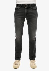 Jeans de mezclilla negros con desvanecimiento sutil, corte recto, cinco bolsillos y un cinturón negro con hebilla de plata. Combinados con zapatillas beige.