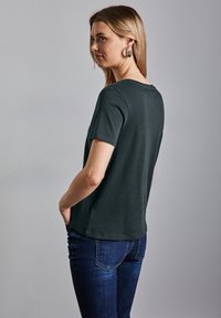 Tmavě zelené tričko s krátkým rukávem a kulatým výstřihem, vyrobené z měkké tkaniny, spárované s tmavě modrými džíny, zobrazené z bočního pohledu.