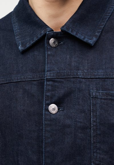 Κοντινή λήψη του γιακά και του μπροστινού μέρους ενός σκούρου μπλε τζιν μπουφάν με μεταλλικά κουμπιά πατιλέτας και εμφανείς λεπτομέρειες ραφής.