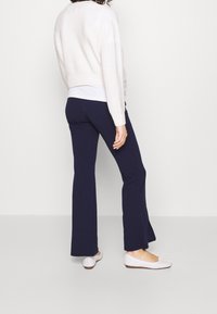 Vaalean beigen värinen kevyesti neulottu villapaita, jossa on nyrkkipunoksella tehtyjä yksityiskohtia, yhdistettynä tummansinisiin leveälahkeisiin housuihin. Mallilla on valkoiset tasapohjaiset kengät, mikä korostaa rentoa asua.