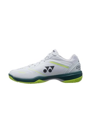 Zapatilla deportiva blanca con suela verde y amarilla, logo negro en el lateral, diseño con cordones y "65" impreso cerca del talón.