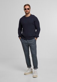 Maglione lavorato a maglia blu navy con un motivo testurizzato, abbinato a pantaloni grigi a sigaretta. Sneakers bianche con accenti beige, modello che indossa occhiali da sole.