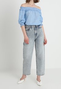 Ljusblå av-axel-blus med volanger, i kombination med högmidjade ljusblå jeans och vita spetsiga mules.