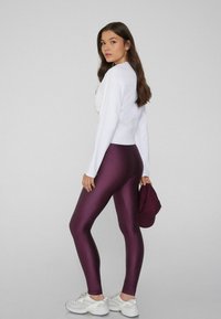 Top bianco a maniche lunghe aderente e leggings bordeaux lucidi a vita alta. Il modello tiene un cappellino bordeaux. Scarpe da ginnastica bianche con accenti argento.