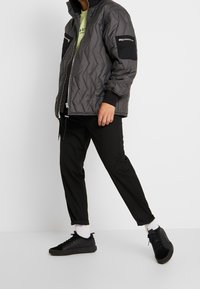 Sort quiltet jakke med høj krave, lynlåslukning foran og sidelommer; parret med sorte bukser og sorte sneakers. Hvide sokker synlige.
