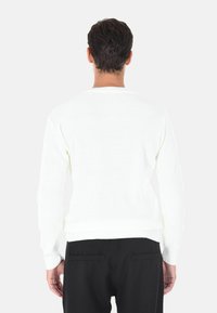 Maglione bianco a maglia con maniche lunghe e texture a coste, con scollo rotondo e vita aderente. Indossato con pantaloni neri.