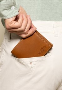 Portafoglio in pelle marrone parzialmente visibile in una tasca di pantaloni chiari. Texture liscia, forma rettangolare, con dettaglio del marchio in rilievo.