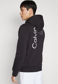 Černá mikina s kapucí s bílým logem "Calvin Klein", se strukturovanou tkaninou a žebrovanými manžetami. Jednoduchý design s uvolněným střihem.