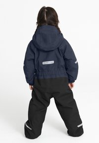 Laivasto takki, jossa on huppu, joustava vyötärö ja heijastavat yksityiskohdat, yhdistettynä mustiin vedenpitäviin housuihin. Yksinkertainen, toiminnallinen muotoilu.