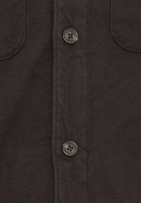 Black fabric shirt with two chest pockets, featuring brown buttons and a textured surface. The button detail includes a logo on one button.