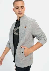 Veste matelassée grise avec un col haut, fermeture éclair, poches avant avec boutons-pression et un petit écusson de marque sur la poitrine.