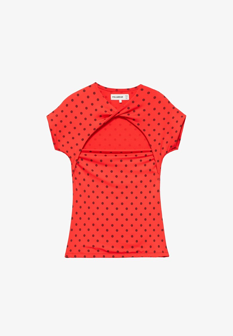 Chemise rouge à manches courtes avec des pois noirs et un détail découpé torsadé au niveau de la poitrine, posée à plat sur un fond blanc.