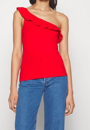 Kvinde iført en rød, enkelt skulders top med en flæsedetalje, kombineret med blå denimjeans, vist fra nakke til hofter.