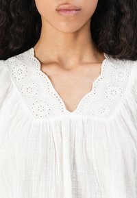 Kvinna som bär en vit blus med scalloped, broderad halsringning och löst, texturerat tyg, som visar hals och övre bröst.