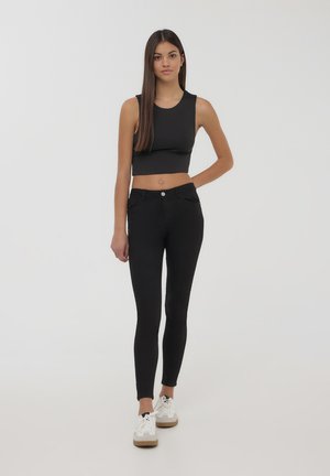 Top corto aderente nero con spalline larghe abbinato a jeans skinny neri a vita alta. Il modello indossa sneakers bianche con suole beige.