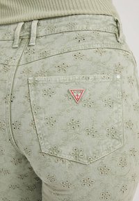 Jeans in denim verde ricamati con motivo floreale, dotati di una tasca posteriore e di un logo a triangolo rosso del marchio "Guess".