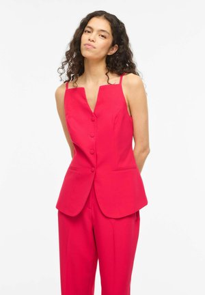 Barna göndör hajú nő áll, piros ujjatlan, gombos mellényt és hozzá illő piros, bő szárú nadrágot visel, egyszínű fehér háttér előtt.