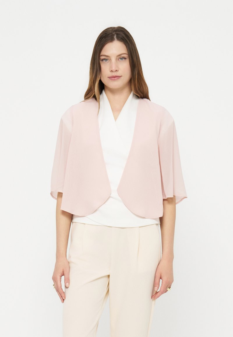 Femme portant un cardigan à manches courtes rose clair ouvert sur un haut blanc et un pantalon crème, debout devant un fond blanc uni.