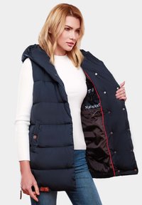 Marineblauer Steppweste mit Kapuze, gestepptem Design, rotem Futter und Druckknopfverschluss. Getragen über einem weißen gerippten Pullover und blauen Jeans.