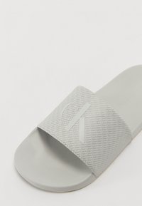 Sandale glissante grise avec une sangle texturée arborant le logo "Calvin Klein" répétitif et un bold "CK", semelle en caoutchouc flexible.
