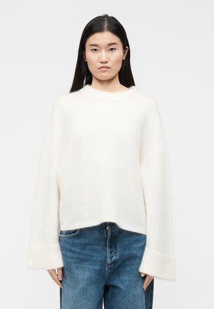 Ženska z dolgimi črnimi lasmi, ki nosi ohlapen bela pulover in modre kavbojke, stoji pred enobarvnim svetlo sivim ozadjem.