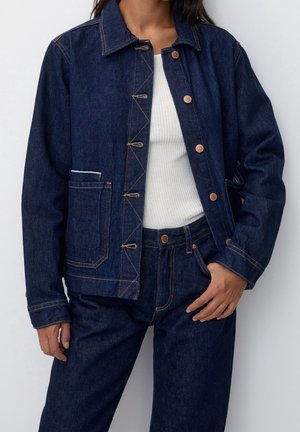 Personne portant une veste en jean bleu foncé et un jean assorti sur une chemise blanche côtelée, avec la veste partiellement boutonnée.